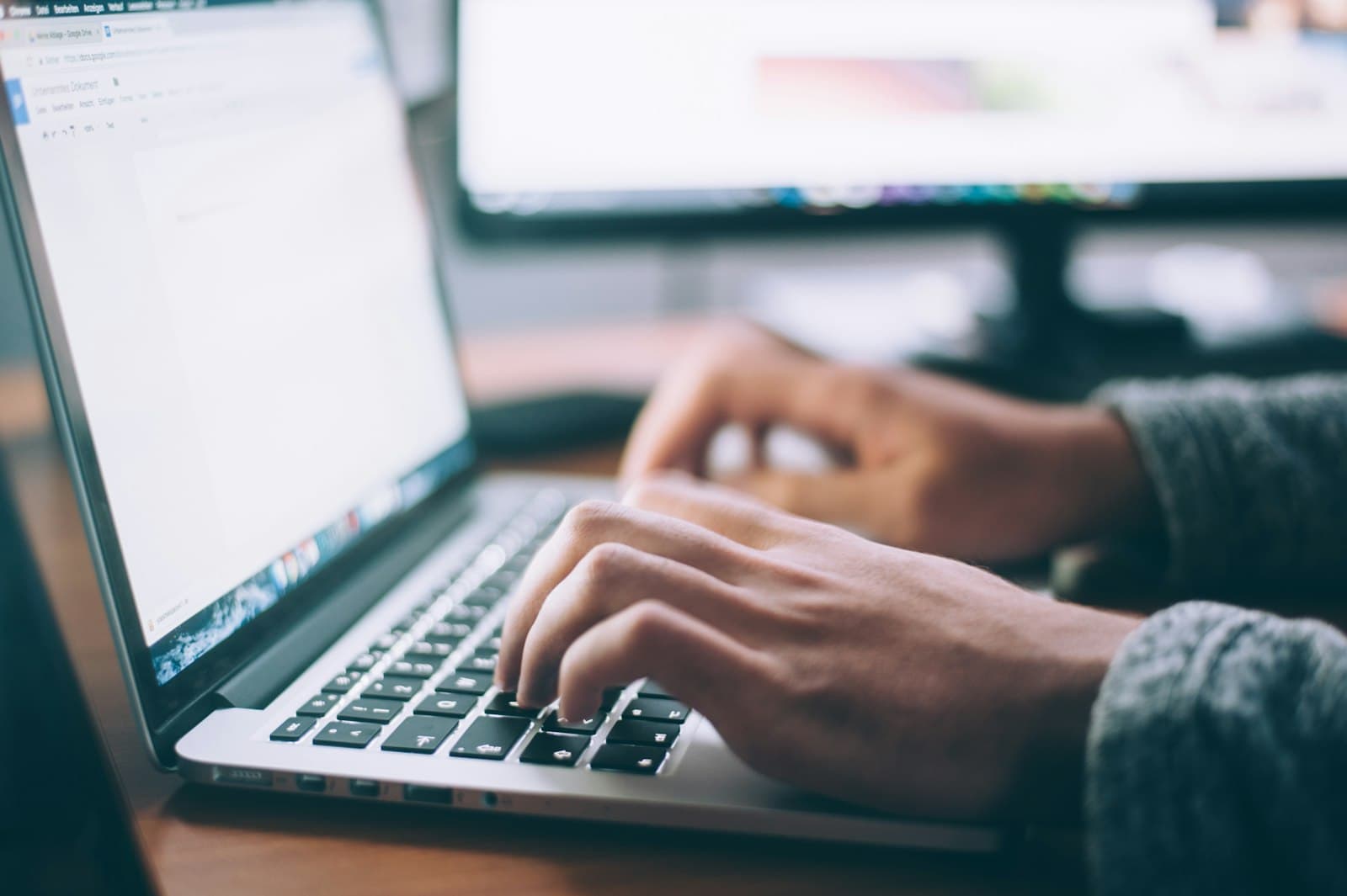 Business owner typing on a laptop at a wooden desk while reviewing documents in natural light (Photo: Unsplash)